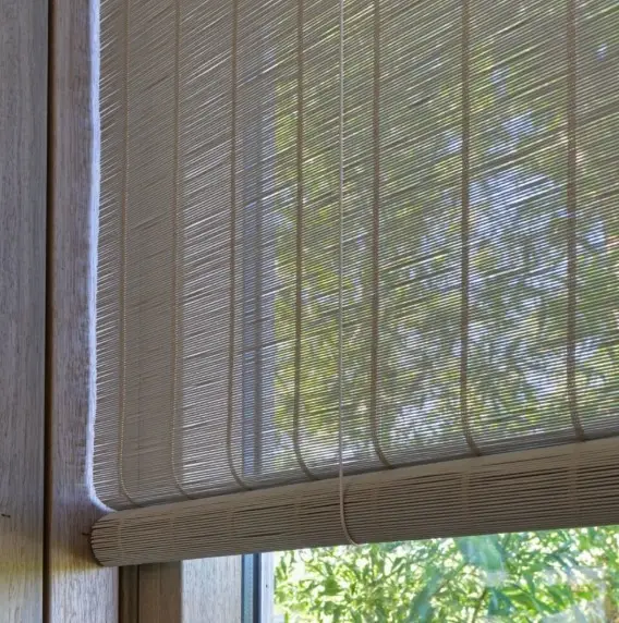 Een close-up van een raam bedekt met een lichtgekleurd, geweven bamboe rolgordijn. Zonlicht filtert door het rolgordijn, waardoor groene boomtakken en bladeren buiten gedeeltelijk zichtbaar worden.