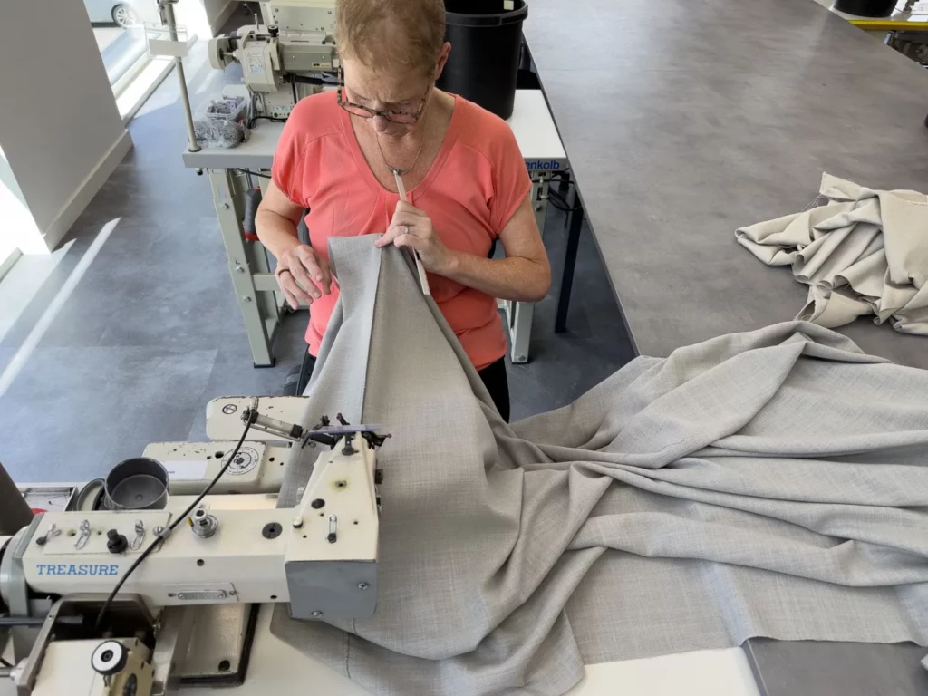 Een vrouw met een bril en een koraalkleurig overhemd naait een groot stuk grijze stof werkt mogelijk aan Raamdecoratie met een naaimachine in een lichte moderne werkruimte met andere machines en stof zichtbaar raamdecoratie rdt concept West Vlaanderen