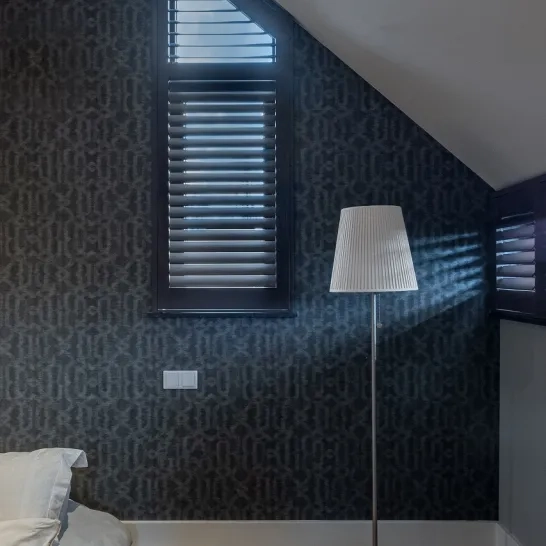 Een moderne slaapkamerhoek met een donkere accentmuur met patroon, een hoge witte staande lamp, een raam met gesloten jaloezieën en een deel van een onopgemaakt bed met wit beddengoed zichtbaar linksonder.