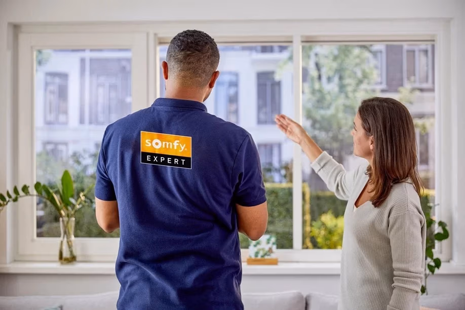 Een vrouw gebaart naar grote ramen terwijl ze praat met een man die een Somfy Expert shirt draagt in een lichte moderne kamer met planten op de vensterbank   RDT concept