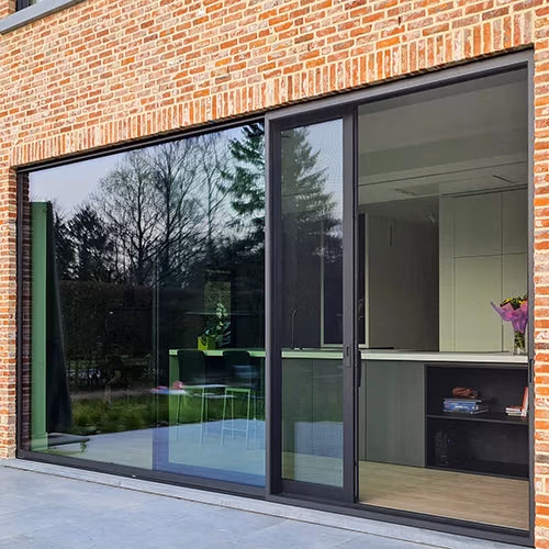 Grote glazen schuifdeur met een zwart frame in een bakstenen muur die bomen en een patio buiten weerspiegelt Binnen is een moderne keuken met witte kastjes en een aanrecht zichtbaar   RDT concept