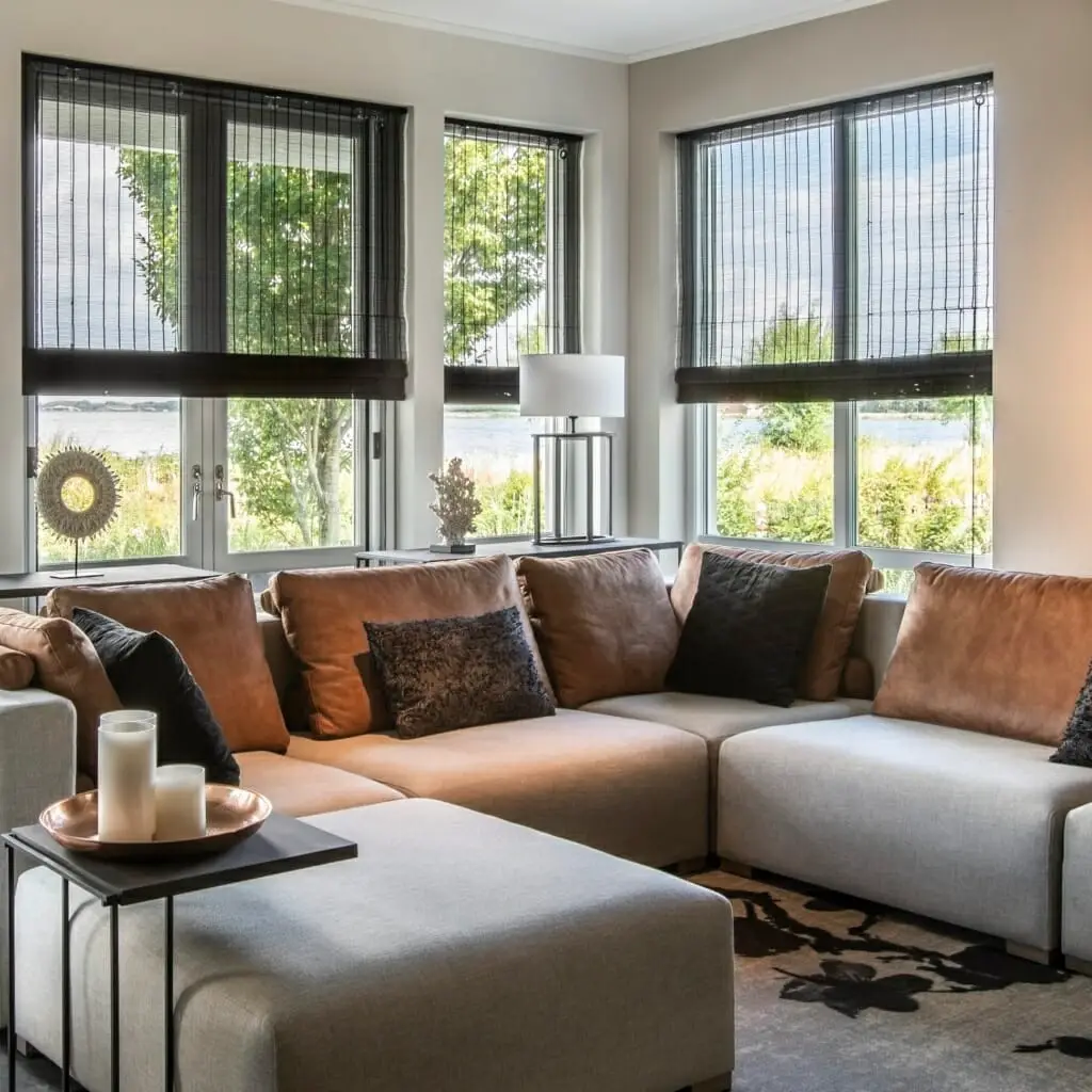 Een moderne woonkamer met grote ramen, een beige sofa met bruine en zwarte kussens, een bijzettafel met kaarsen en uitzicht op groen en water buiten. Geweven houten jaloezieën bedekken de ramen gedeeltelijk.
