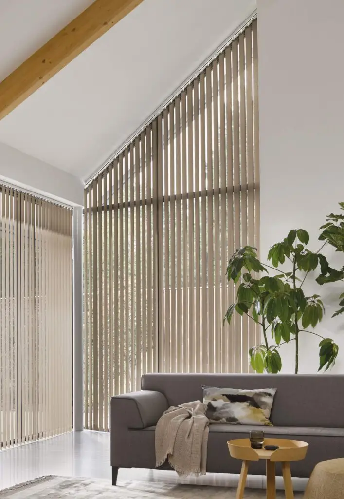 VVB_1612_q7QRIVo2e16d0bafill-1500x2173 - RDT Concept Een moderne woonkamer met hoge schuine ramen bekleed met beige lamellen een grijze bank met een kussen met patroon een houten salontafel en een grote plant in pot Natuurlijk licht vult de ruimte RDT Concept